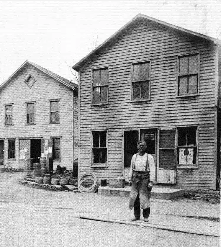 Photo with caption: John Sintz Senior is pictured here in the early 1900s. Blacksmith and repair work was done in the building to the left. Both buildings still stand in the middle of town on US 52, practically unrecognizable from their original form.